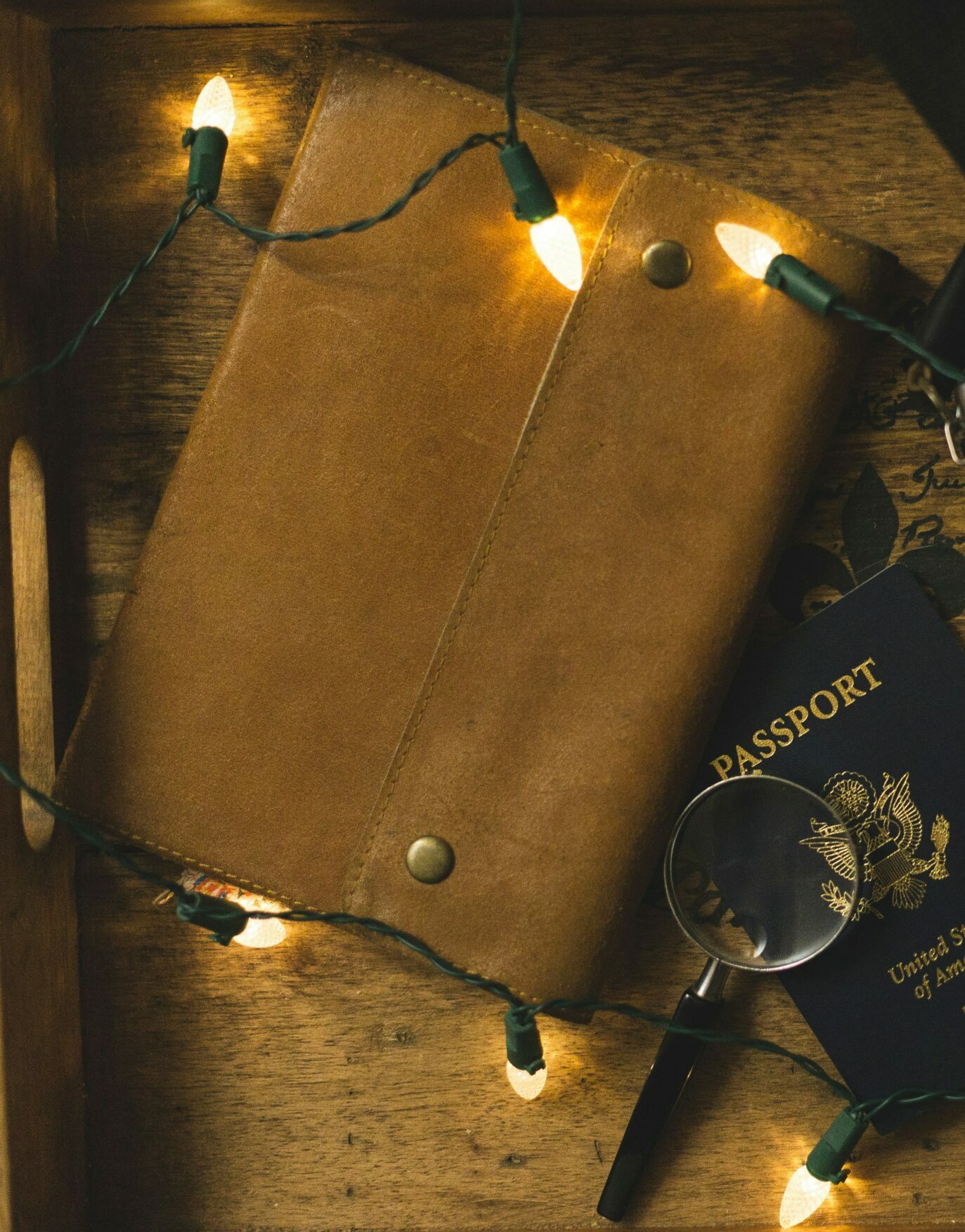 Brown leather notebook next to magnifying class and passport on wooden surface with white Christmas lights