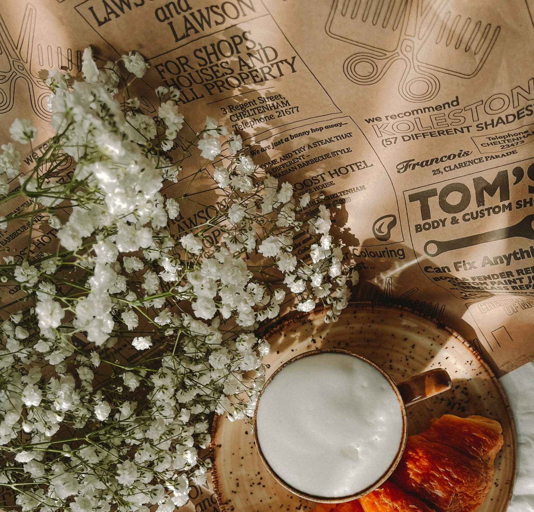 White baby's breath on brown newspaper next to wooden plate with cup of frothed milk and a croissant