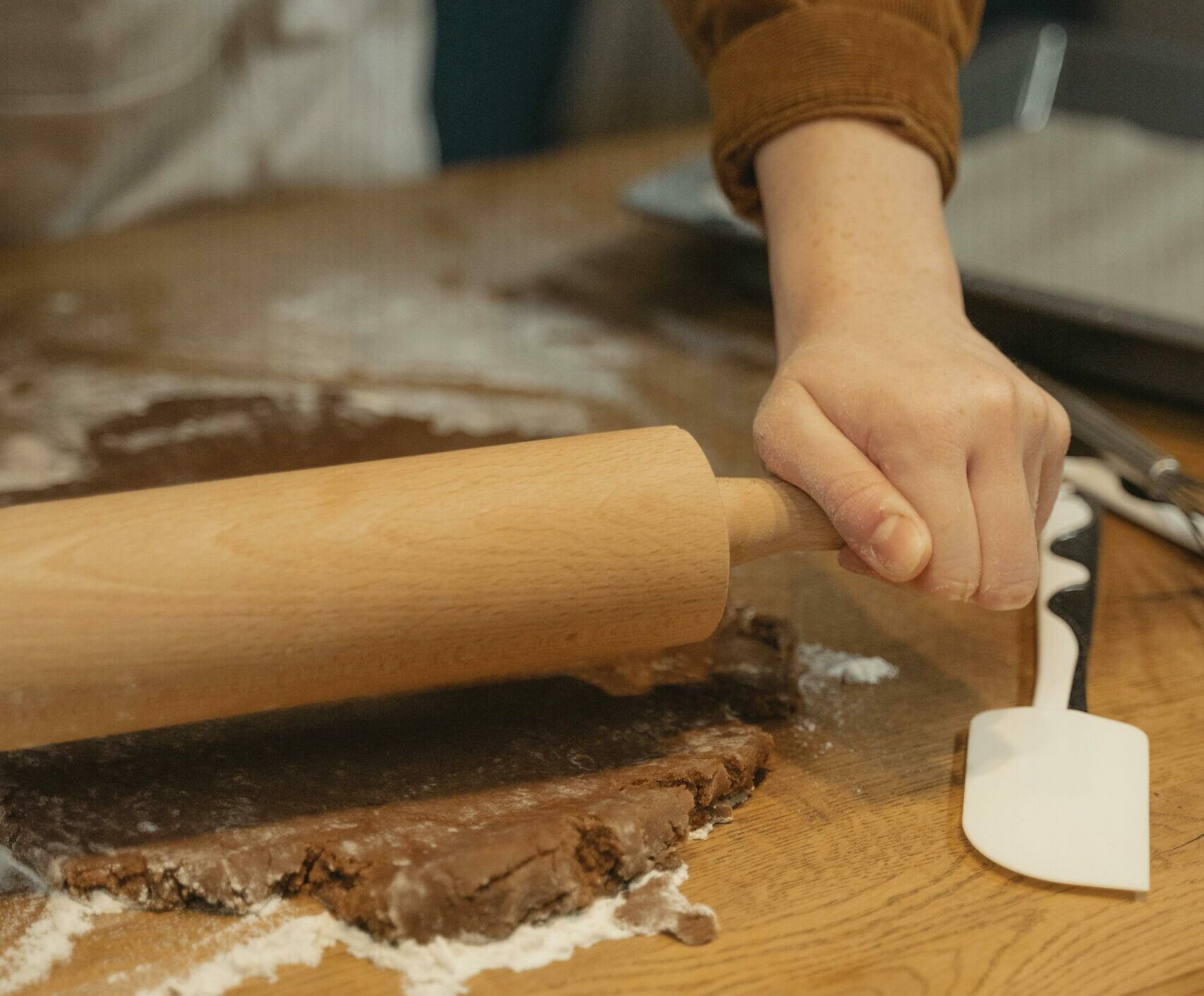 Person rolling out brown dough next to white rubber spatula on wooden counter